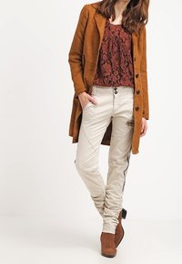 Brązowy zamszowy długi płaszcz, burgundowa koronkowa bluzka, jasnobeżowe dopasowane spodnie z czarnymi bocznymi paskami oraz brązowe botki na kostkę. Formalna kreacja.