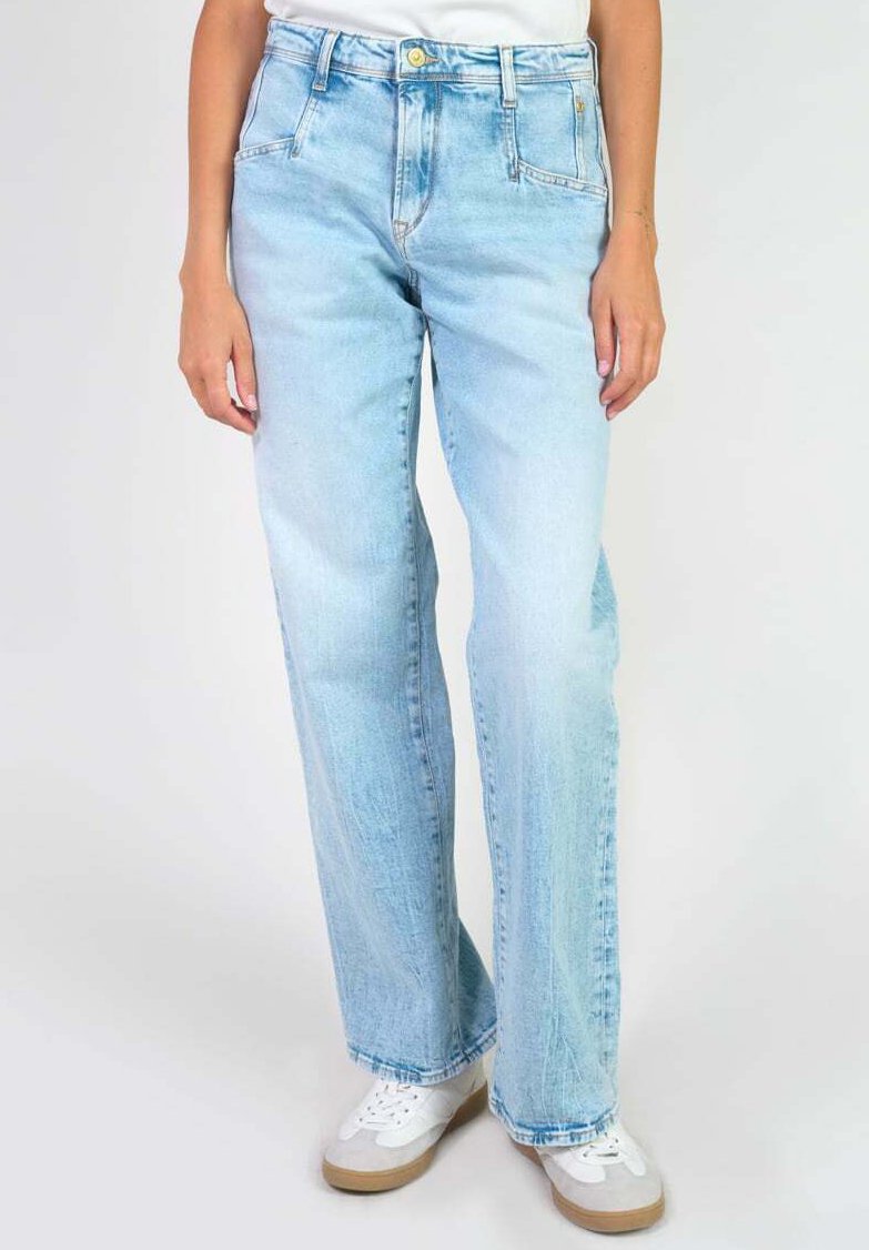Jean en denim bleu clair à jambe large avec une taille haute et une texture délavée. Équipé d'une fermeture à bouton et de poches latérales. Associé à des baskets blanches.