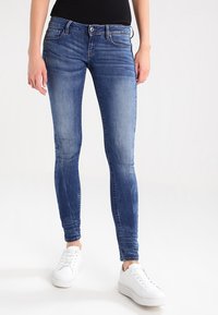 Uskeh skinny traperice u srednje plavom denimu, s izblijedjelim završetkom, pet džepova i petljama za remen. Uparene s bijelim tenisicama.