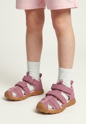 Børnebens ben iført hvide sokker og lyserøde lukkede sandaler med brune såler, parret med lyserøde shorts mod en ensfarvet baggrund.