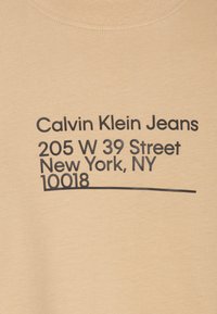 Béžové bavlněné tričko s tučným černým textem: "Calvin Klein Jeans 205 W 39 Street New York, NY 10018" se jednoduchým podtržením.
