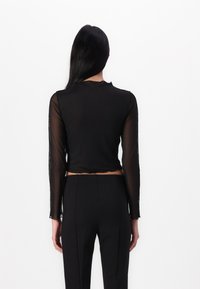 Črn skrajšan top z dolgimi prosojnimi rokavi, izrezom z valovitim robom in teksturirano tkanino, v kombinaciji z krojenimi črnimi hlačami.