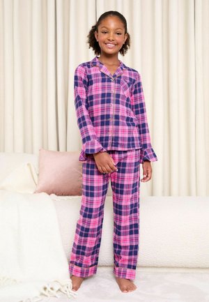 Fată tânără zâmbitoare, stând desculță într-un pijama în carouri roz și albastru închis, într-o cameră luminoasă moale cu draperii și perne în nuanțe neutre.