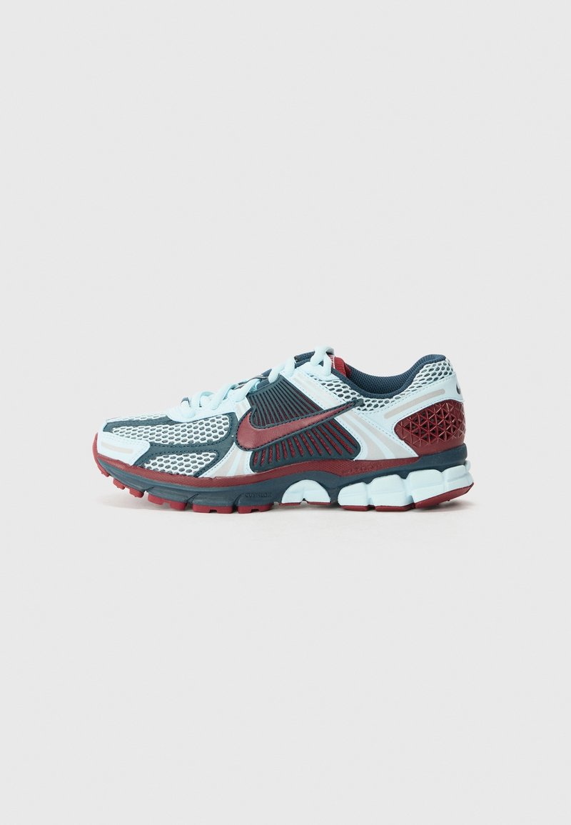 Chaussure de course Nike blanche, bleue et rouge avec un design en mesh, semelle amortissante visible et logo swoosh rouge sur fond gris.