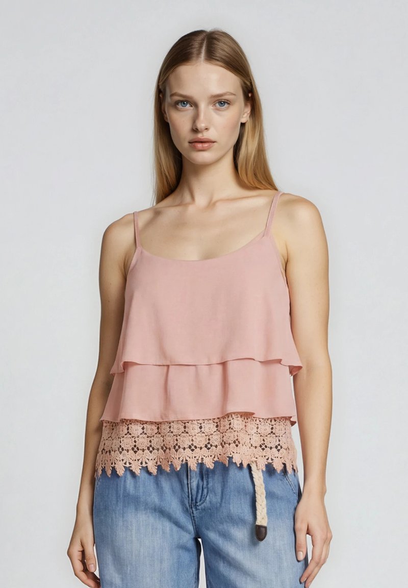Femme blonde portant un haut rose sans manches superposé avec bordure en dentelle et un jean en denim bleu sur un fond blanc uni.