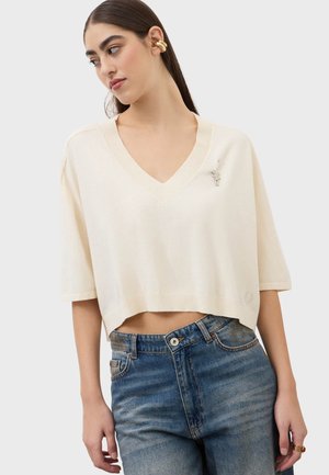 Femme portant un pull court crème à col en V avec des manches mi-longues, un jean bleu, un manchon d'oreille doré et une broche argentée sur le pull.