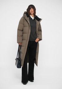 Doudoune de couleur marron terne avec une silhouette longue, dotée d'une capuche et d'un design matelassé carré. Associée à un pull sombre et à un pantalon noir.