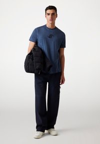 Marineblå struktureret t-shirt med logo, sort pufferjakke draperet over armen, og mørke cargo-bukser med lynlåse og lommer. Hvide sneakers.