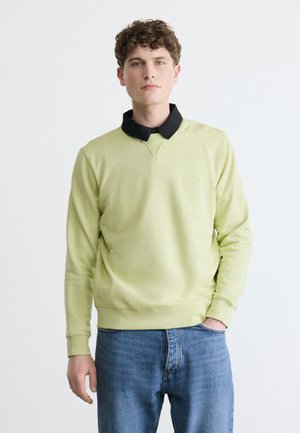 Noor mees lokkis juustega, kandes helerohelist dressipluusi üle musta kraega pluusi ja siniseid teksaseid, seisab ühe käega taskus.