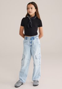 Must ristkülikukujuline t-särk, helerohelised lahtised püksid küljetaskutega ja mustvalgete mustritega libisevad kingad. Lihtne, vabaaja riietus.