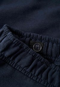 Granatowy materiał z elastycznym paskiem w talii i zebraną fakturą. Posiada przezroczysty guzik z czterema otworami, przymocowany do paska.