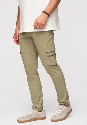 Mężczyzna w oliwkowych spodniach typu cargo z klapami i kieszeniami na zamek, białych trampkach i białej koszulce, stojący na jednolitym tle.