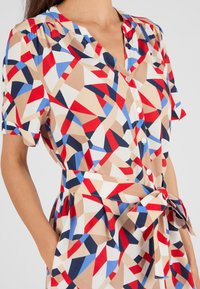Korte mouwen jurk met een geometrisch patroon in rood, blauw en beige. Gemaakt van gladde stof met een knopen sluiting aan de front en een ceintuur in de taille.