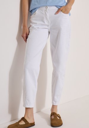 Persoon met een witte broek, blauw overhemd en bruine suède gespschoenen staat met één hand in de zak tegen een lichte achtergrond.