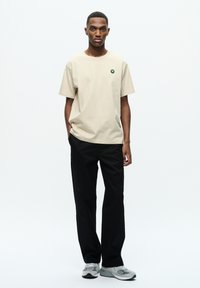 Beige bomuld t-shirt med et lille grønt logo på brystet, parret med sorte bukser og grå sportssko. Simpelt design og afslappet pasform.