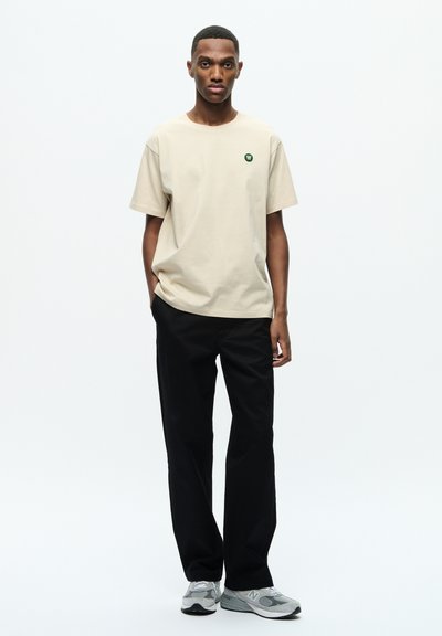 Beige Baumwoll-T-Shirt mit einem kleinen grünen Logo auf der Brust, kombiniert mit schwarzen Hosen und grauen Sportschuhen. Einfaches Design und entspannter Schnitt.