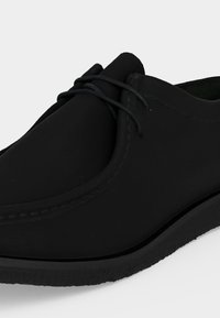 Chaussure décontractée en cuir noir lacée, bout arrondi, semelle épaisse et coutures visibles sur le dessus, gros plan sur fond blanc.