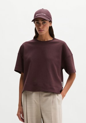 Frau trägt ein lockeres, dunkelviolettes Kurzarmoberteil, beige Hose und eine passende dunkelviolette Baseballmütze mit weißem Text.