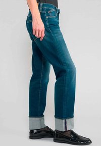 Jeans en denim bleu profond avec une coupe droite. Présentent des ourlets roulés avec un tissu contrastant. Portés avec des mocassins noirs brillants.