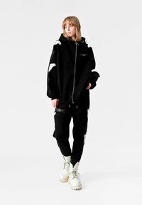 Persona vistiendo una sudadera negra de gran tamaño y pantalones a juego con diseños de alas blancas, de pie contra un fondo blanco, usando botas blancas.