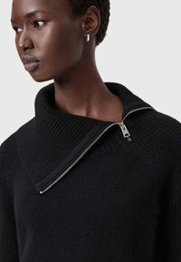 Μαύρο πλεκτό πουλόβερ με φαρδύ ριμπ λαιμόκοψη και μεταλλική λεπτομέρεια στο φερμουάρ στον λαιμό. Η υφή φαίνεται μαλακή και άνετη.