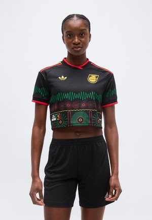 Jeune femme portant un maillot de football noir de la Jamaïque, court, avec des motifs rouges, verts et jaunes, et un short noir Adidas, debout devant un fond blanc.