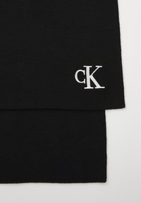 Svart strikket skjerf med en myk tekstur, med en hvit brodert "cK" logo på den ene enden. Enkel, minimalistisk design.