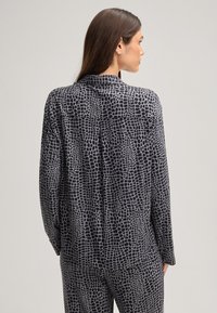 Zwarte blouse met lange mouwen en een grijs krokodillenhuidpatroon. Voorzien van een kraag, knoopsluiting aan de achterkant en een ontspannen pasvorm met zichtbaar merklogo.