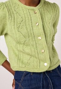 Detailní záběr na osobu, která má na sobě světle zelený pletený kardigan se zlatými knoflíky a tmavě modré džíny s oranžovým prošíváním.