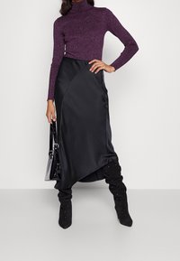 Černá saténová asymetrická sukně spárovaná s těsně přiléhajícím fialovým rolákem s žebrováním. Modelka nosí zdobené černé kozačky nad kolena a drží kabelku.