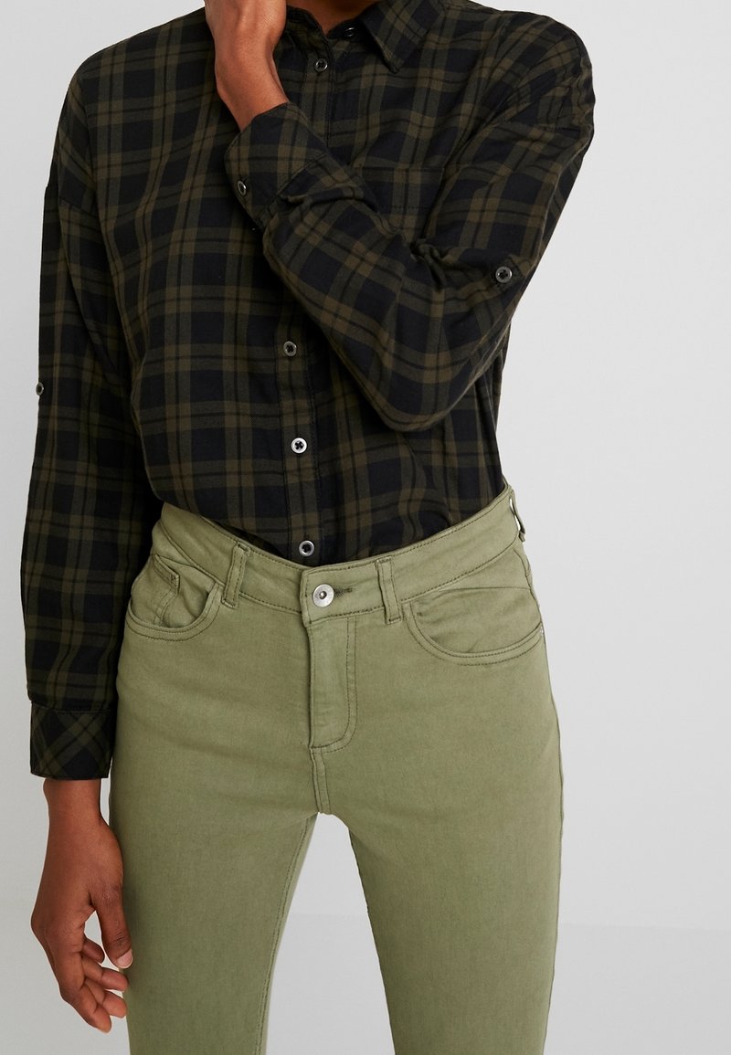 Chemise à col boutonné en tissu tartan noir et vert, assortie à un jean en coton slim-fit vert olive avec poches avant et fermeture à bouton.