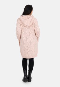 Un long cardigan rose en tricot avec une capuche, présentant un motif de tricot torsadé et des poignets côtelés, porté sur des leggings noirs et de lourdes bottes noires.