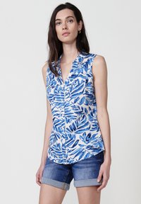 La camicia senza maniche con bottoni presenta un motivo a foglia blu e bianco su un tessuto leggero. Abbinata a pantaloni shorts in denim con orli arrotolati.