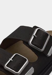 Κοντινό πλάνο μαύρων λουριών σανδαλιού με δύο ασημένιες ορθογώνιες πόρπες και μπεζ πάτο που διακρίνεται από κάτω.