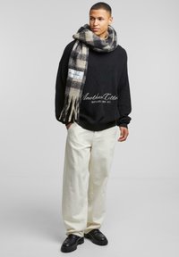Maglione nero a maglia con dettagli a costine, pantaloni crema a gamba larga e una sciarpa in plaid grigia con bordi frangiati. Scarpe nere chunky completano il look.