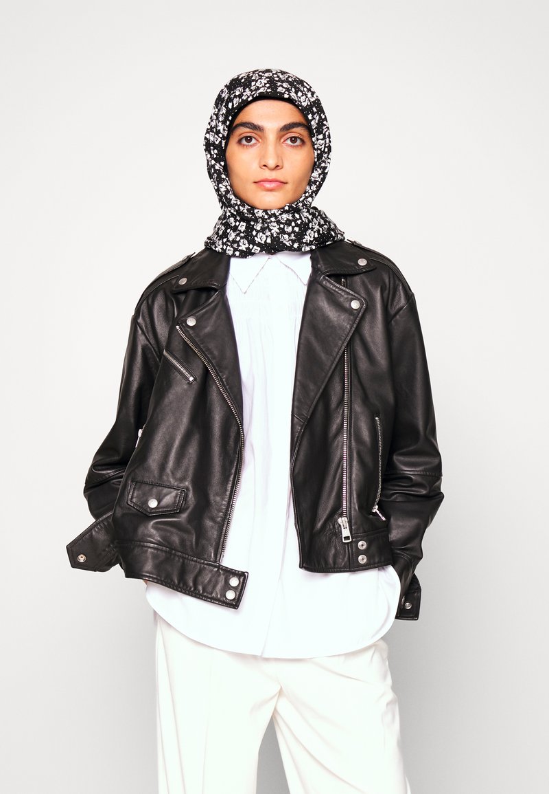 Casaco de couro preto estilo biker com fechos e botões prateados, combinado com uma blusa branca e um lenço com padrão floral em preto e branco.