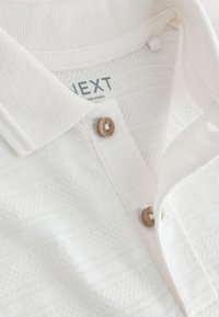 Fehér mintás galléros póló fából készült gombokkal és a gallér belsejében látható "NEXT" felirattal, Indiában készült.