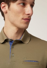 Olivově zelené polo tričko s modrým kostkovaným vnitřním límcem a detailem na kapse; obsahuje dva knoflíky a vyšitý logo psa na hrudi.