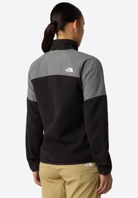 Černá fleecová bunda s šedou horní částí. Vybavena vysokým límcem a logem na zadní straně. Měkká textura, neformální design a dlouhé rukávy.