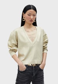 Maglione dorato chiaro a V con maniche ampie e orlo a costine. Abbinato a jeans scuri. La modella ha i capelli lunghi e indossa grandi orecchini geometrici.