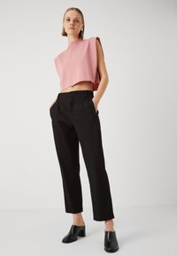 Roze cropped top met een hoge hals, gecombineerd met zwarte plooibroeken en zwarte hoge hakken, tegen een neutrale achtergrond.