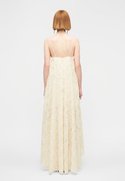 Μακρύ ελεφαντόδοντο φόρεμα με ροή στη σιλουέτα, φτιαγμένο από υφή υφάσματος με λουλουδένια σχέδια. Λεπτές τιράντες και χαμηλή πλάτη.