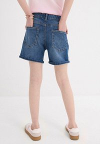 Shorts in denim blu con orli arrotolati e due tasche posteriori. Indossati con scarpe chiare. Texture liscia e design casual.