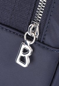 Sac en tissu bleu marine avec fermeture éclair argentée et un tirant en métal distinctif comportant une lettre "B" découpée. Texture lisse avec des coutures visibles.