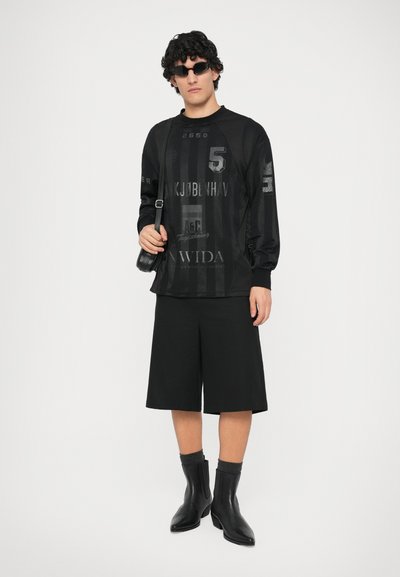 Sudadera gráfica negra con patrones de texturas variadas, combinada con pantalones cortos negros y botas hasta el tobillo. Los accesorios incluyen gafas de sol negras pequeñas y un bolso de hombro.