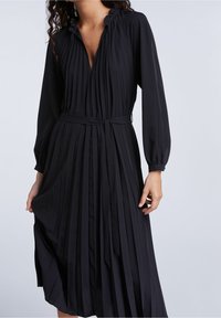 Robe noire plissée avec des manches longues, un col froncé et une taille ceinturée. Le tissu semble lisse avec des plis verticaux bien marqués.