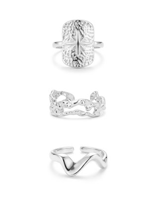 Tres anillos de plata: el superior rectangular con diseño de estrella, el del medio banda orgánica calada, el inferior anillo abierto liso con forma de ola, todos sobre fondo blanco.