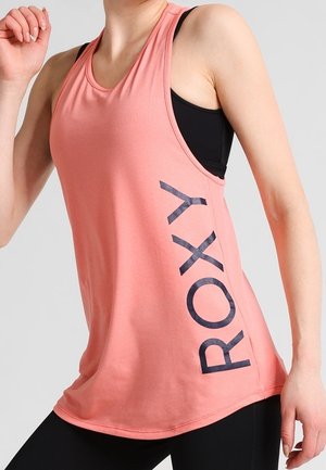 Vrouw draagt een mouwloos koraalroze tanktop met zwarte "ROXY" tekst aan de zijkant, gecombineerd met zwarte leggings.