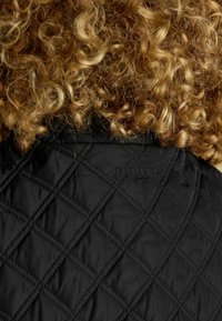 Lockiges, hellbraunes Haar über einer schwarzen, gesteppte Jacke mit "Joules" bestickt am hinteren Kragen.