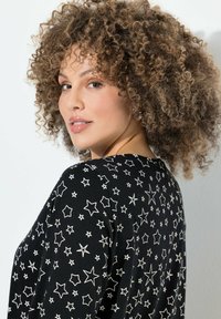 Schwarzes Stoffoberteil mit weißen Sterne-Mustern, runder Ausschnitt und lockerer Passform. Die Haare sind in sanften, voluminösen Locken gestylt.